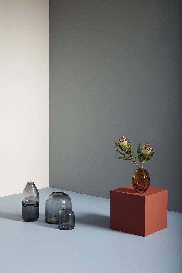 En moderne stilleben-komposition med en HÜBSCH - Dome Vaser, der understøtter en vase med blomster, flankeret af to glasskulpturer, sat mod en tofarvet grå og hvid baggrund.