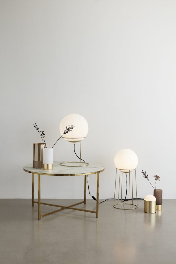 Et minimalistisk interiør med et rundt bord med en gennemskinnelig grøn top og guldben, ledsaget af tre globusbordlamper og små dekorative vaser med kviste. Baggrunden er en neutral grå væg