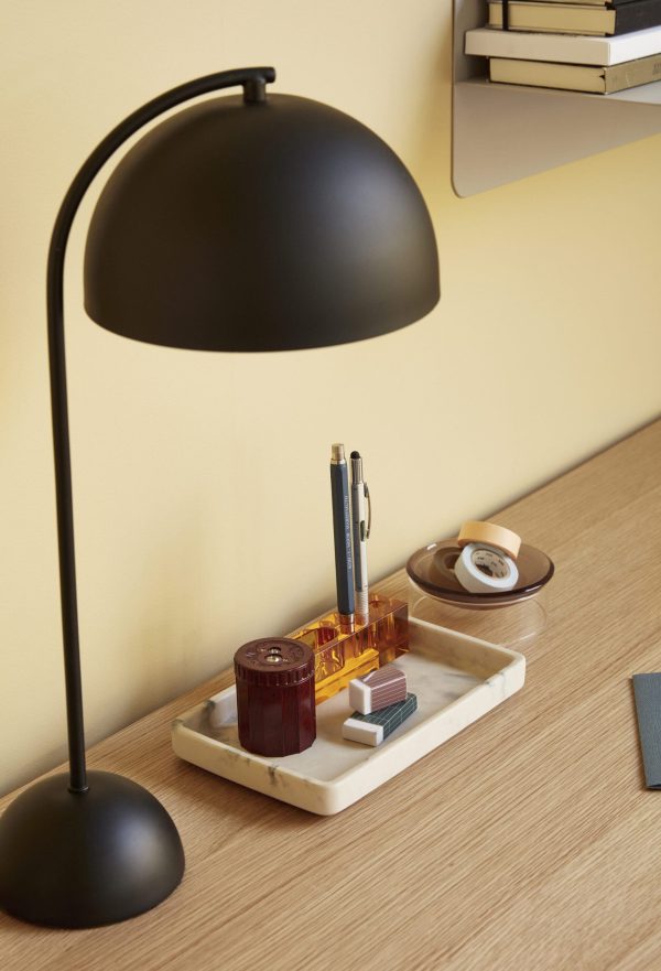 En sort skrivebordslampe med buet hals oplyser en ryddelig skrivebordsopsætning med en hvid bakke med papirvarer, ved siden af stablede bøger på en lys træoverflade mod en gul væg. Denne scene fremhæver