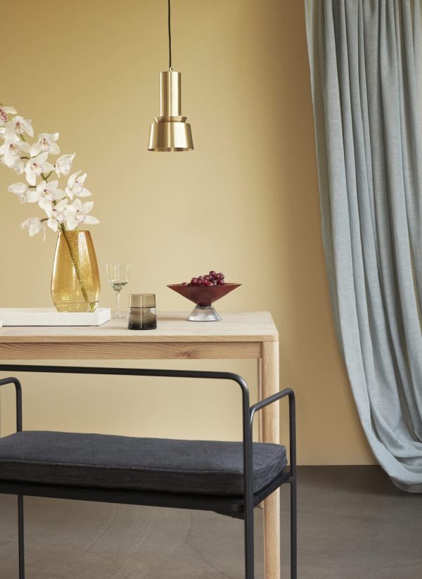 En moderne spiseplads med et træbord, en sort bænk med en pude og en Tomorrowsdesign pendel ovenover. Bordet er prydet med en vase med hvide blomster og en skål med