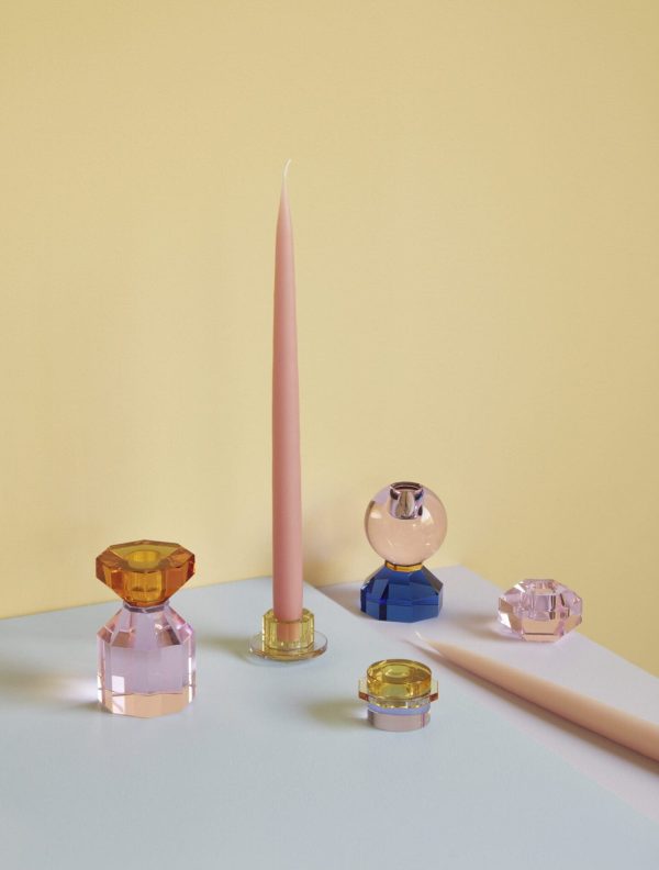 Et lyserødt stearinlys står oprejst blandt flere farverige lysestager i glas på gul baggrund og skaber et roligt og velordnet indretningsarrangement.