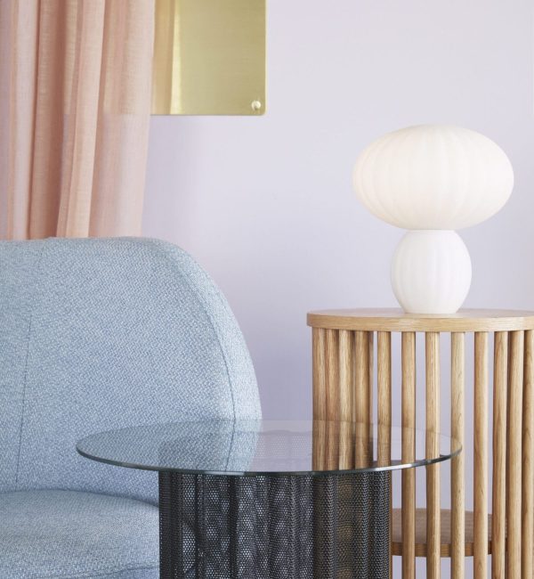 Et moderne stuehjørne med en blå tekstureret lænestol, et sidebord i træ med en sfærisk hvid lampe fra Tomorrowsdesign og et rundt sofabord i glas mod en pastelrosa væg med