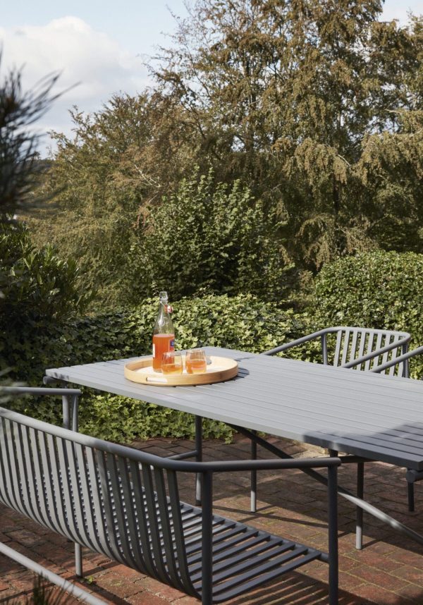 En rolig udendørs indstilling med et designergråt havebord med to matchende stole, en bakke med en flaske og to glas orange drik, omgivet af frodige grønne områder under klar himmel.