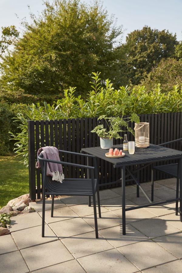 En udendørs terrasse med et sort spisebord med matchende stole, omgivet af frodige grønne områder. Bordet er prydet med en plante, stearinlys og en bakke HÜBSCH - Aster Drikkeglas, under solrige himmelstrøg.