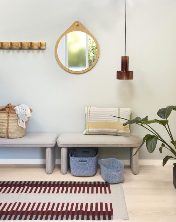 En moderne, minimalistisk entré med to grå designerbænke med hynder, et rundt spejl, pendellamper og et stribet tæppe. En vævet kurv, blå opbevaringskurve og en plante tilføjer dekorative detaljer