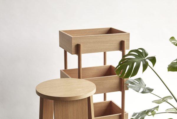 En minimalistisk træplantestativ med flere niveauer, der viser en blanding af åbne hylder og skuffer, inkorporerer Tomorrowsdesign på en neutral baggrund med en delvis udsigt til et grønt monstera-blad.