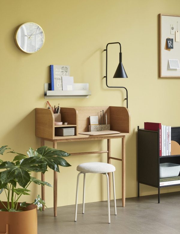 Et hjemmekontoropsætning med et træskrivebord og skammel mod en sennepsgul væg. Skrivebordet har hylder med bøger og papirvarer, ledsaget af en Tomorrowsdesign vægmonteret lampe og ur, næste