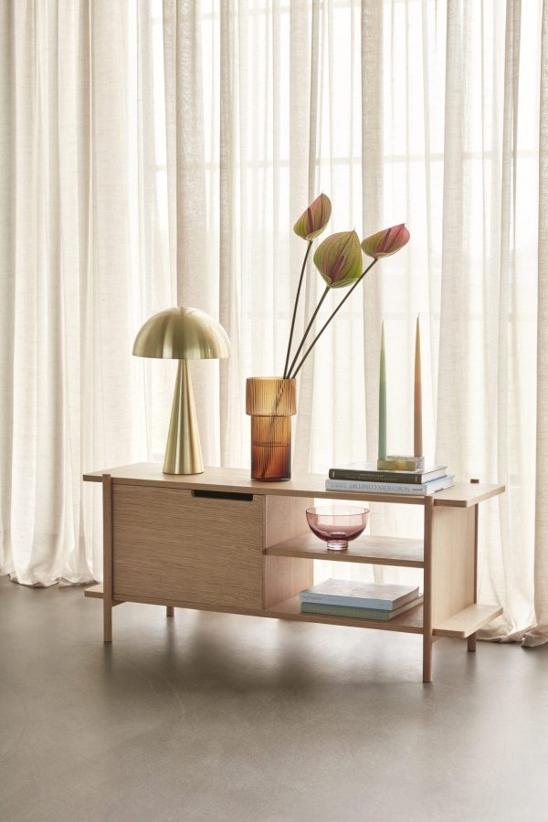 En moderne stue med en skænk af egetræ med dekorative elementer, herunder en metallisk lampe, HÜBSCH - Ripple Vase og en glasskål. Gennemsigtige gardiner tillader naturligt lys at fylde rummet.