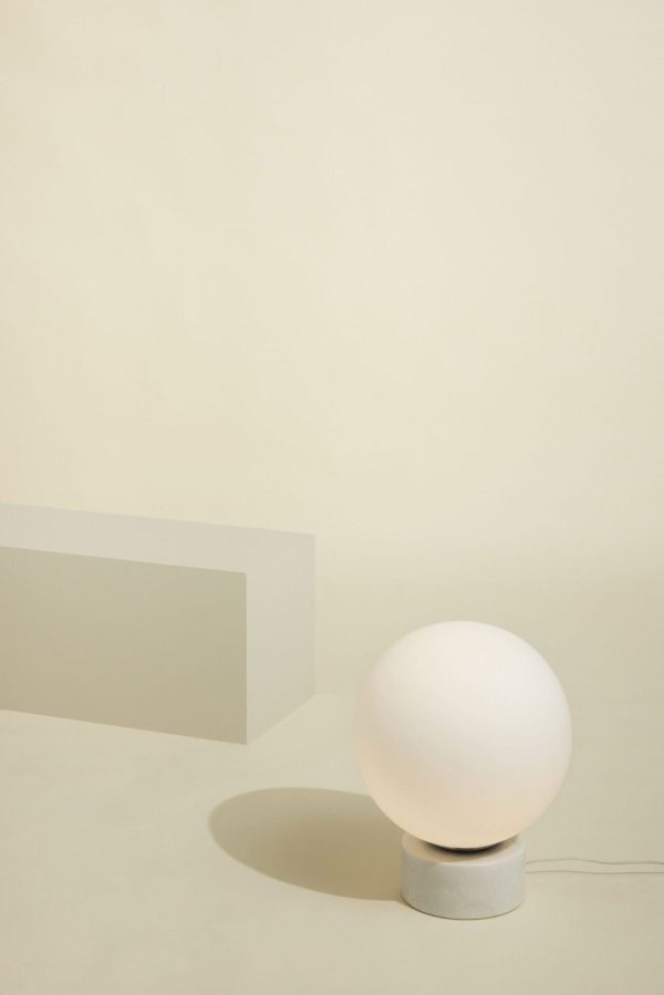 En minimalistisk komposition med en HÜBSCH - Sphere Lampe på en cylindrisk grå base, sat mod en lys beige baggrund med en rektangulær blok til venstre. lampen kaster en blød skygge.
