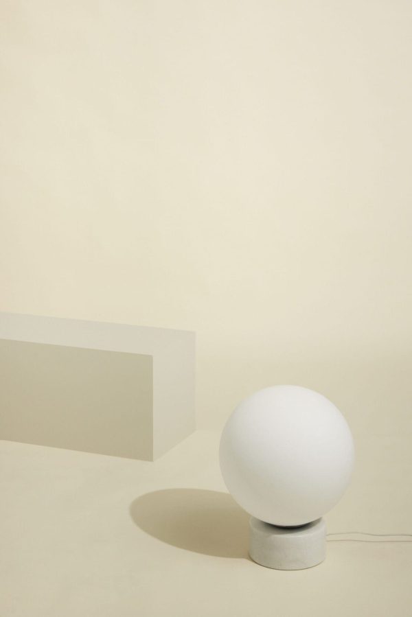 Et minimalistisk setup med en HÜBSCH - Sphere Lampe på et lille gråt stativ, placeret ved siden af en enkel, rektangulær blok mod en lys beige baggrund.