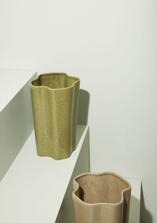 En keramisk vase med en tekstureret, plettet finish står på en hvid trappeplatform og kaster delvist en skygge på overfladen. Vasen har en unik asymmetrisk overkant og inkarnerer et innovativt design