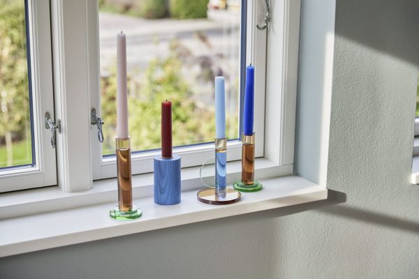 Tre farverige stearinlys i designerholdere i en vindueskarm, med udsigt til en trækantet gade udenfor. Lysene er røde, hvide og blå, hver i en unik gennemsigtig base.