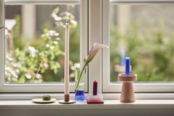 Et fredfyldt vindueskarm-arrangement med en calla-lilje i en glasvase, flankeret af stearinlys og farverige geometrisk-formede Tomorrows-designgenstande, med en solbeskinnet have synlig gennem vinduet.