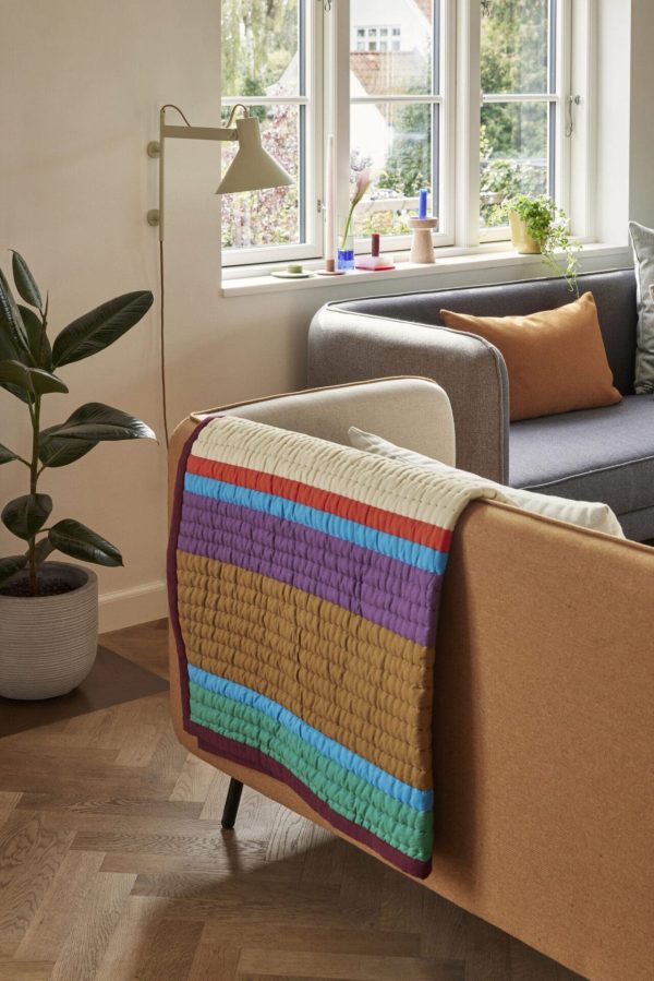 Et hyggeligt stuehjørne med en grå sofa med et farverigt vævet tæppe på, en stor plante i en krukke og et vindue med udsigt til det grønne. En gulvlampe og små stearinlys