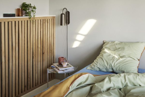 Et moderne soveværelseshjørne med en træspaltevæg, et sidebord med bøger og en kop og en seng med grønt og blåt sengetøj. En vægmonteret lampe kaster blødt sollys hen over rummet,