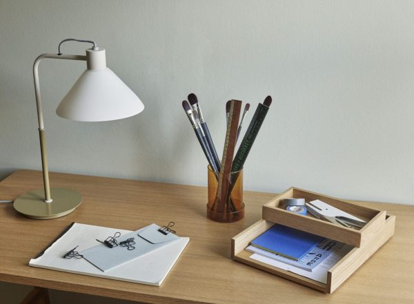 Et pænt organiseret skrivebord med en hvid lampe, en åben notesbog med designskitser, en træholder med malerpensler og en lille træbakke med kontorartikler.