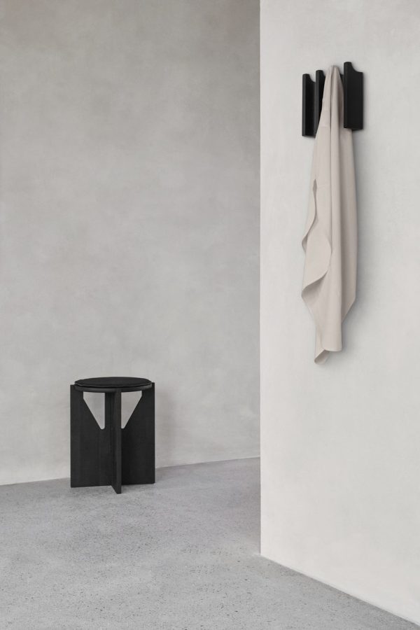 Et minimalistisk værelse med gråt gulv og væg, med en sort skammel til venstre og en beige frakke, der hænger på sorte kroge til højre. Den overordnede atmosfære er moderne og skarp og inkarnerer