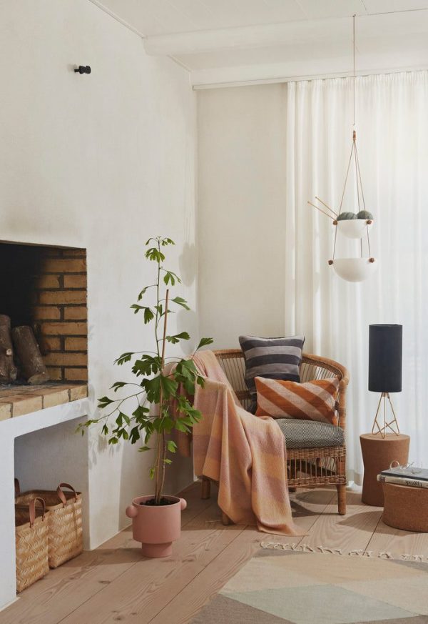 Et hyggeligt stuehjørne med en kurvestol med hynder og puder, en hvid pejs med brænde, en høj potteplante og blød belysning fra en unik overheadlampe. Lysgardiner og
