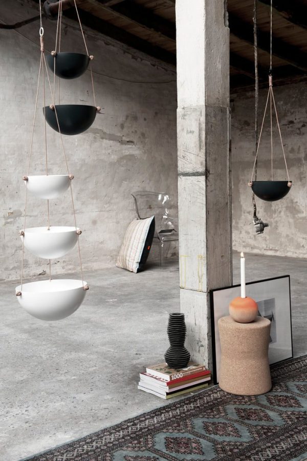 Moderne plantekasser, der hænger i snore i et minimalistisk betoninteriør med et mønstret tæppe, en stak bøger, indrammet kunst og dekorative genstande, der viser en stilfuld blanding af urban og bo