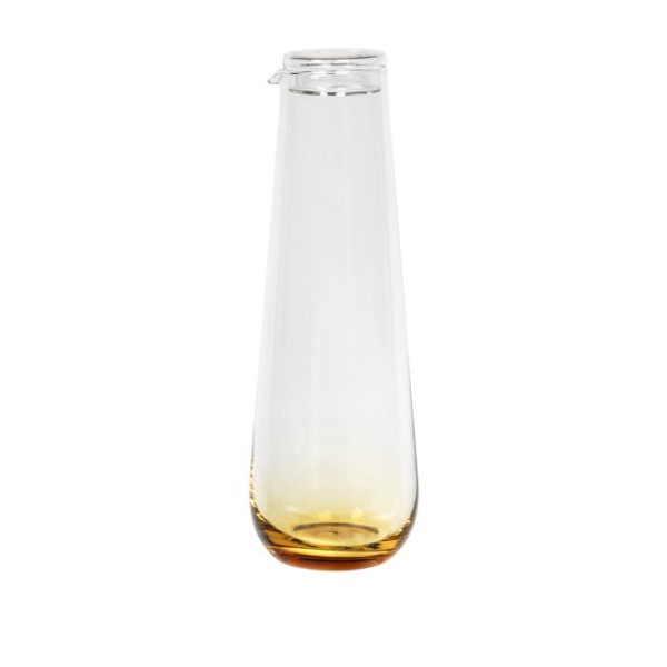 En klar glasflaske med en lille mængde gul væske i bunden, stående oprejst på en hvid baggrund. BROSTE COPENHAGEN - 'Amber' Mundblæst Kande har en smal hals og et hængslet glaslåg.
