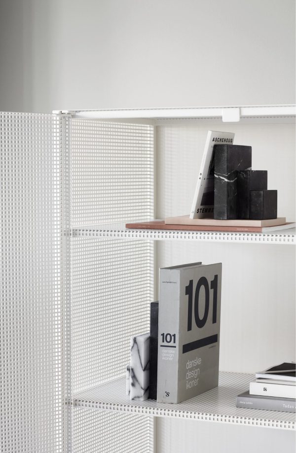 En minimalistisk hvid metal bogreol med perforerede sider, der viser et pænt arrangement af bøger primært fokuseret på design og arkitektur fra "TomorrowsDesign.