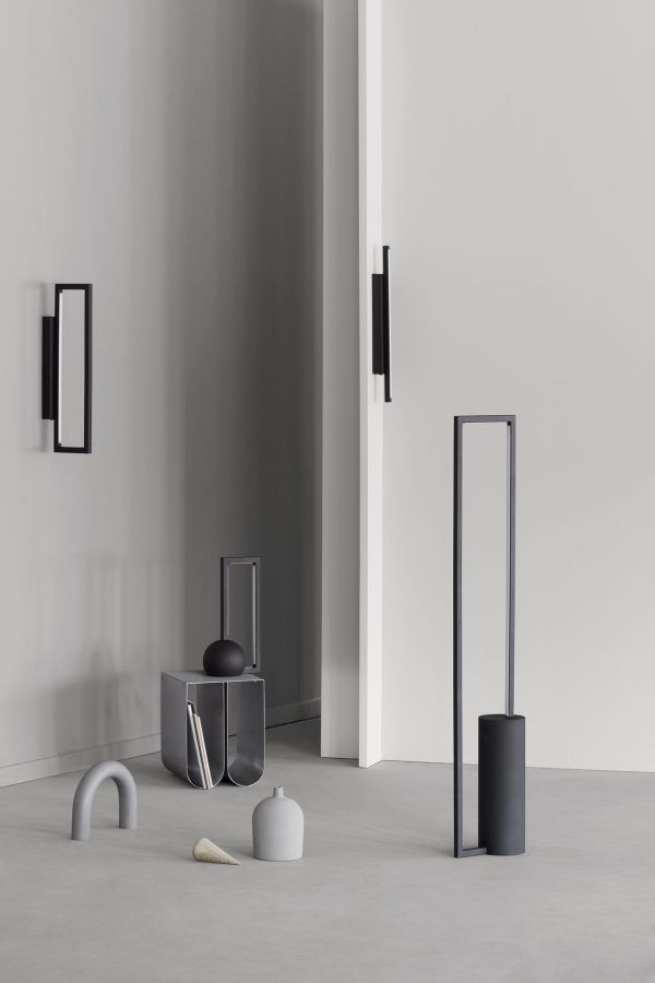 Et minimalistisk rum dekoreret med abstrakte skulpturer og geometriske former i grå og sorte nuancer, med en høj, slank fritstående skulptur og mindre bordpladeobjekter. Neutrale vægge og glatte gulve fremhæver det moderne design