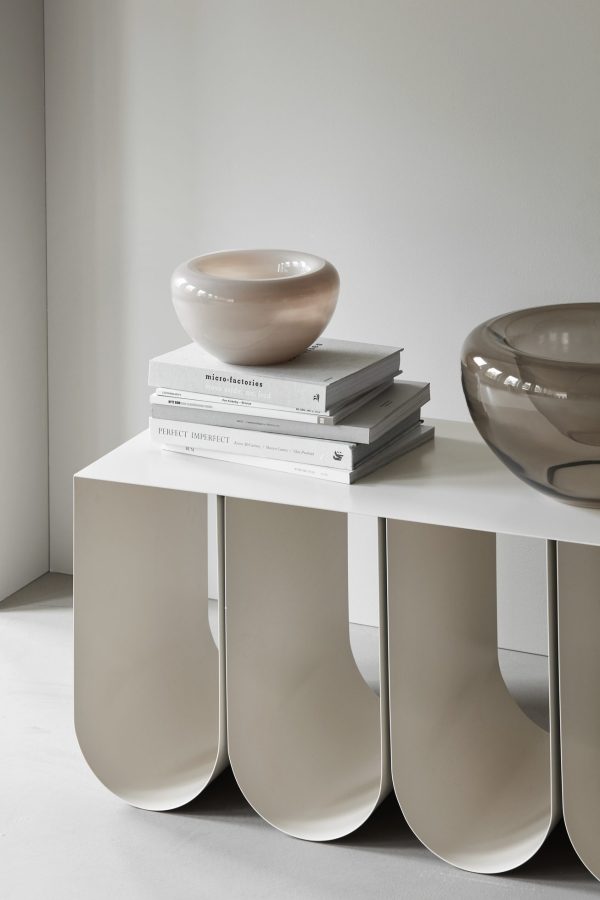 En minimalistisk indretningsscene byder på en hvid modulær bænk med bøger med titlen "perfekt ufuldkommen" og en beige skål ved siden af en større brun skål på et gråt gulv. Neutrale toner dominerer indstillingen.
