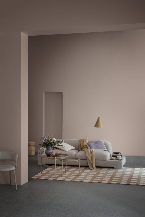 En minimalistisk stue med bløde lyserøde vægge, med en lysegrå sofa prydet med puder og puder, et lille sofabord med bøger, en lampe og planter, på baggrund af en stribet baggrund