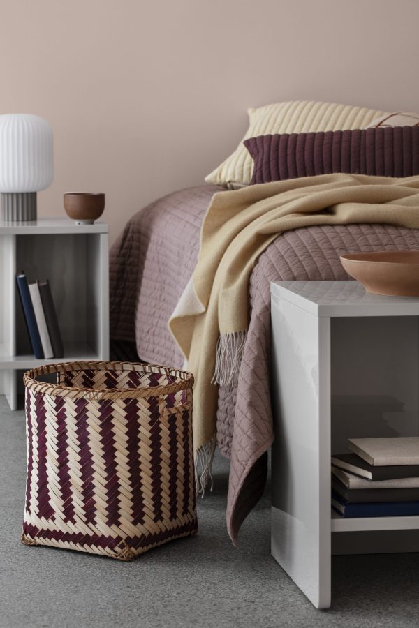 Et hyggeligt soveværelseshjørne med en pænt redt seng med lilla dyne og beige plader, en vævet kurv, et moderne natbord med bøger fra TomorrowsDesign og en lille bordlampe mod en