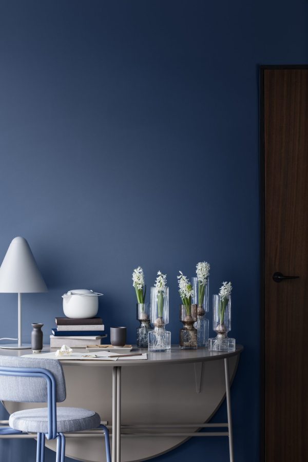 Et studiehjørne i minimalistisk stil med et hvidt skrivebord med en række bøger, stearinlys, en lampe og flere glasvaser med hvide blomster mod en dybblå væg. En trædør og en