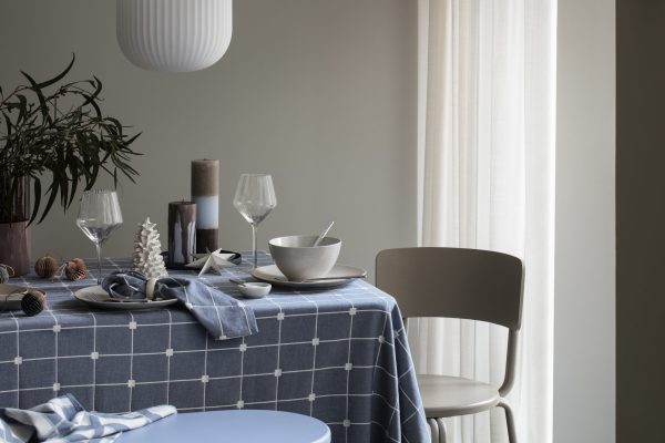 Et pænt dækket spisebord med moderne indretning i et roligt, naturligt oplyst rum. Funktionerne omfatter en blåternet dug, elegant glas, keramiske fade og dekorative genstande som stearinlys og en lille