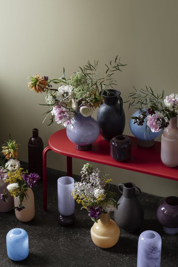 Flere farverige BROSTE COPENHAGEN - 'Mari' Vaser i forskellige former og størrelser arrangeret på et rødt bord med et udvalg af friske blomster i hver, sat op mod en afdæmpet grøn væg.