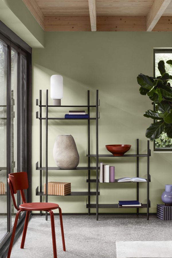 Et moderne værelse med en stor grøn væg og synlige loftsbjælker i træ. Der er sorte minimalistiske hylder med bøger og forskellige vaser. En rød stol, der fremviser dristigt interiørdesign, er placeret