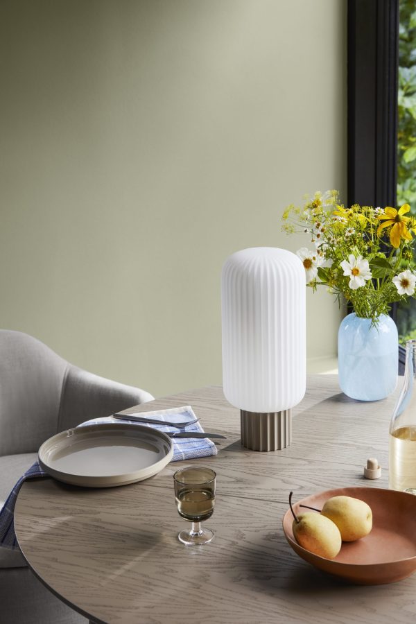 Moderne spisebordsdækning med stilfuld ribbet lampe, hvid tallerken, glas vand, træskål med pærer og vase med gule blomster. Der er en grå sofa og udsigt