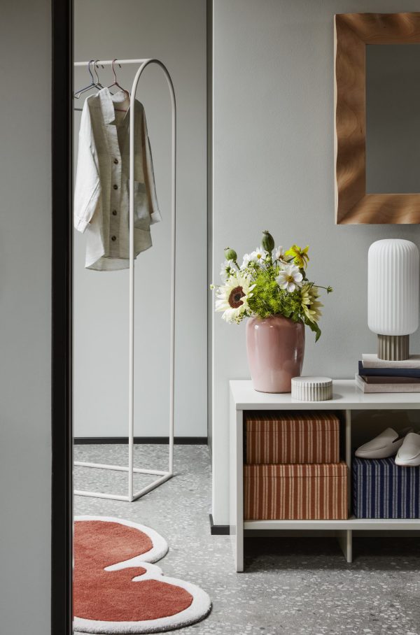 En stilfuld entré med en BROSTE COPENHAGEN - 'Platon' S Marmor Dåse med en hængende hvid jakke, et konsolbord i træ med bøger og en hvid lampe, et hjerteformet tæppe og et spejl på væggen. En vase med blomster tilføjer et strejf af farve.