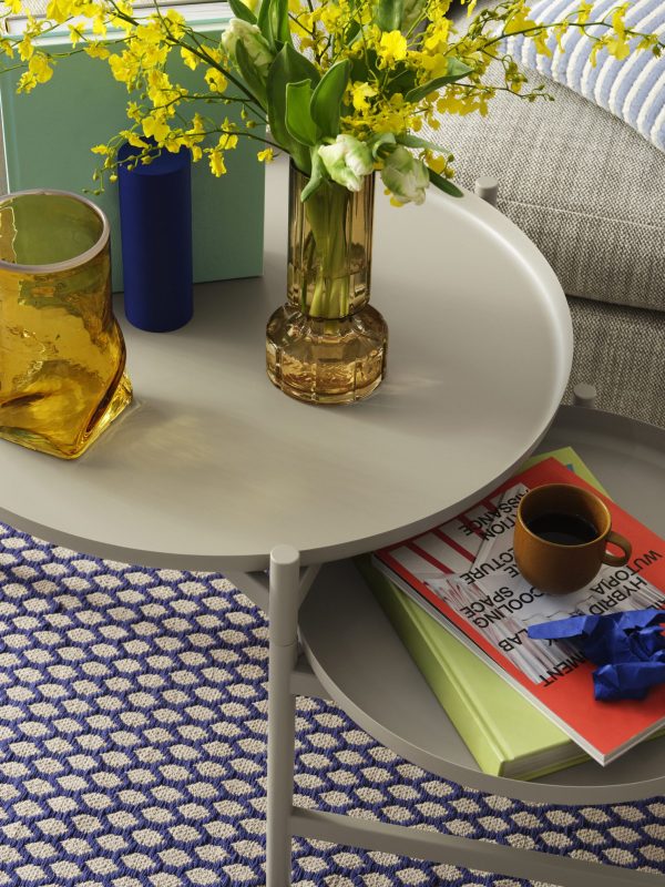 Et pænt anrettet sofabord med bøger, et gult og grønt blomsterarrangement i en glasvase, en kop kaffe og farverige pyntegenstande. Indstillingen omfatter designmøbler såsom en blå