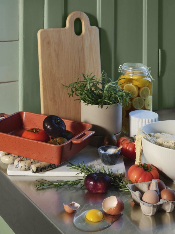 En køkkenbord med friske ingredienser: æg, tomater, krydderurter og citroner. der er en keramikgryde med en plante, en træ BROSTE COPENHAGEN - 'Olina' F.eks. Skærebræt og et bagefad. blødt naturligt lys tilføjer varme.