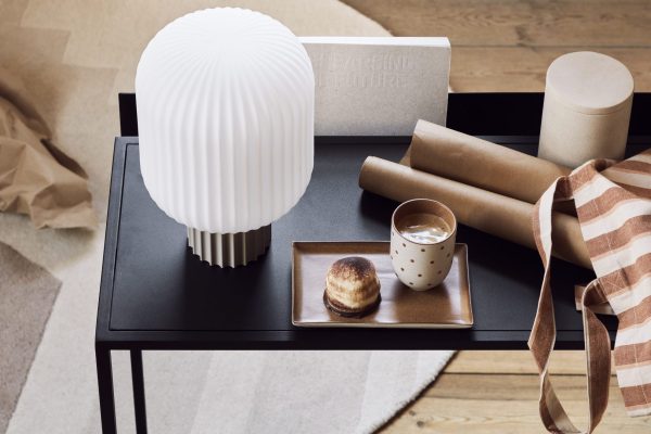 Et stilfuldt anrettet lille sort bord med en stablet bog mærket "undervisningsglæde", en cylindrisk hvid lampe med interiørdesign, en kop latte og en muffin på en