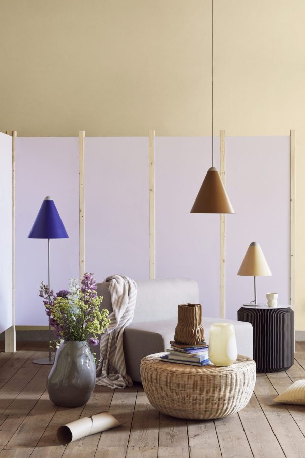 Et hyggeligt interiør med en BROSTE COPENHAGEN - 'Pam' Bord, en stor vase med blomster og diverse lamper i forskellige former. baggrunden omfatter skillevægsskærme i træ med pastel-lilla paneler.
