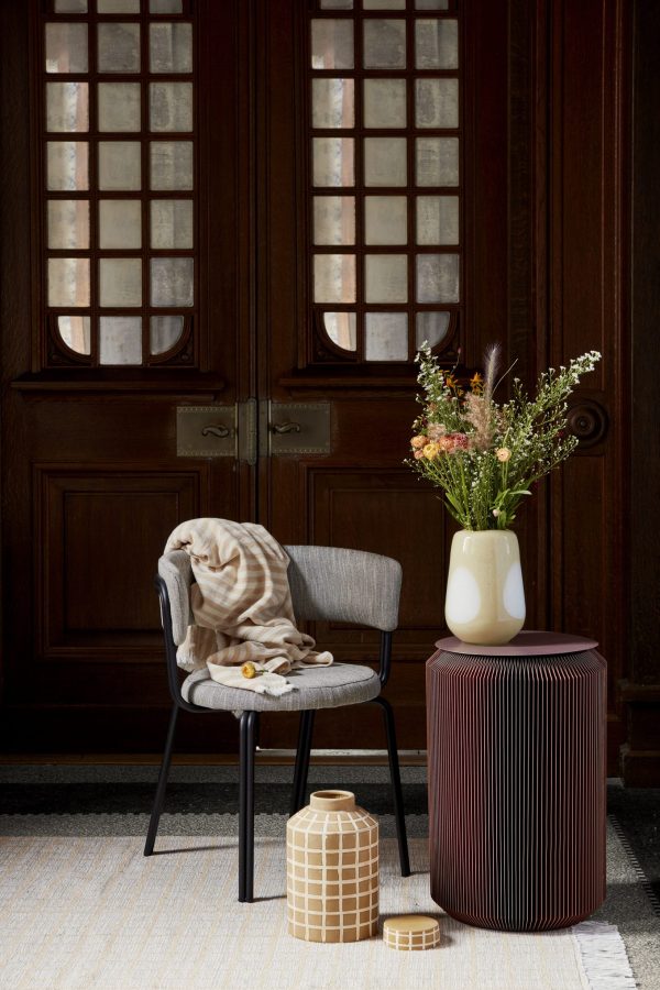 Et hyggehjørne med en elegant grå stol med et draperet beige tæppe, en høj hvid vase med efterårsblomster på en ribbet mørkerød skammel og en lille mønstret vase sat mod rigt