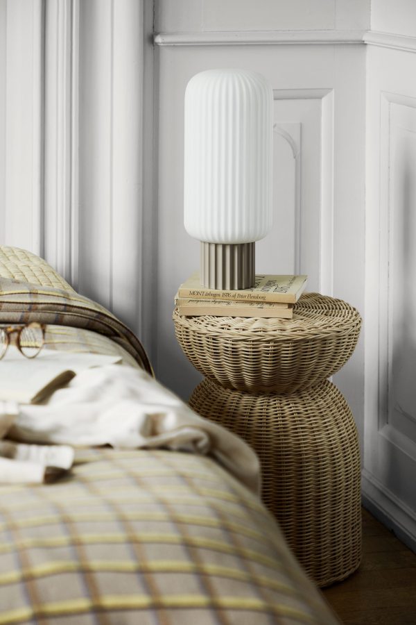 En moderne hvid lampe sidder på toppen af en stak natborde i rattan ved siden af en pænt redet seng med stribet sengetøj, placeret i et værelse med klassiske hvide panelvægge med elegant designmøbler.
