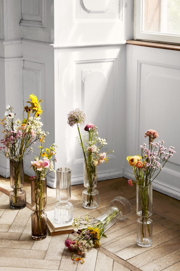 Adskillige klare glasvaser i forskellige højder indeholdende diverse vilde blomster er arrangeret på et sildebenstrægulv nær en hvid panelvæg og store solbeskinnede vinduer, der komplementerer interiørdesignet