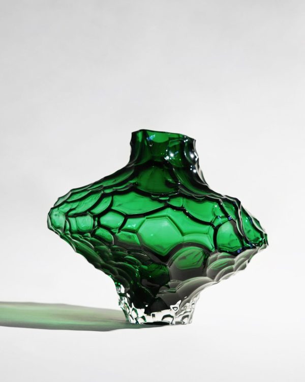 Et nærbillede af en grøn, indviklet glasskulptur, der ligner en smeltende form, vist mod en almindelig lys baggrund. Skulpturen fremviser en levende, blank finish og tekstureret overflade med et strejf af