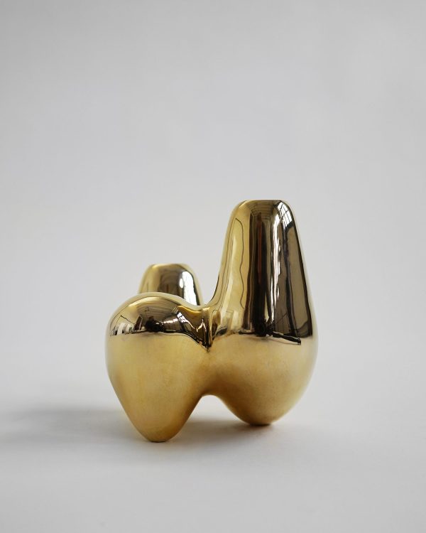 En blank, guldkeramisk skulptur med et glat, abstrakt og løgformet design står på en neutral grå baggrund. Skulpturen har en reflekterende overflade med bløde kurver.