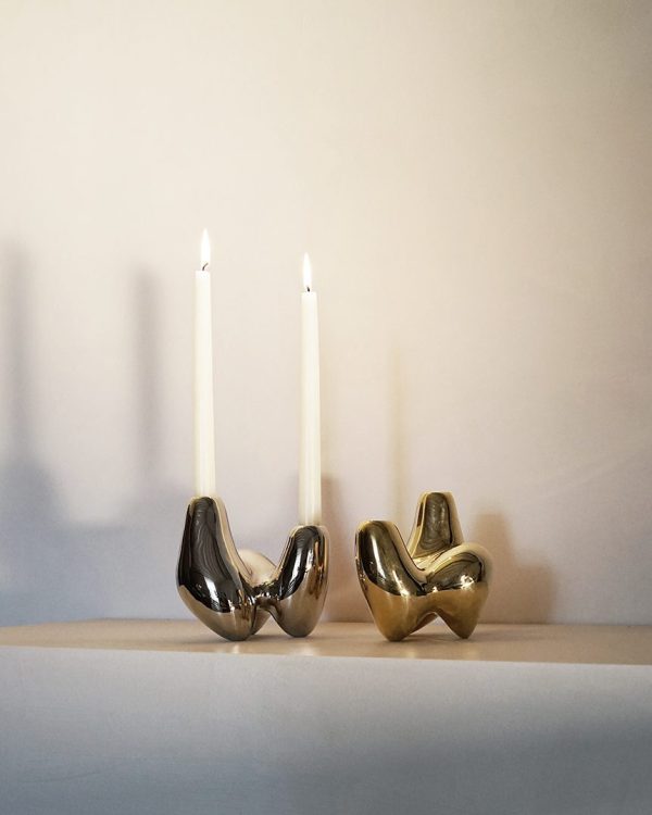 To unikke lysestager, en sølv og en guld, hver formet som et krøllet stykke papir fra Tomorrowsdesign-kollektionen, der holder hvide tilspidsede lys på en let hylde mod en neutral