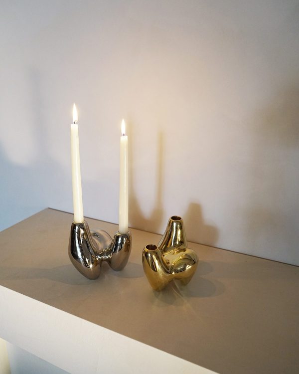 To tændte stearinlys i skinnende, metalliske holdere formet som abstrakte, kurvede skulpturer på en lys beige hylde mod en blødt oplyst hvid væg, der inkarnerer essensen af Tomorrowsdesign.