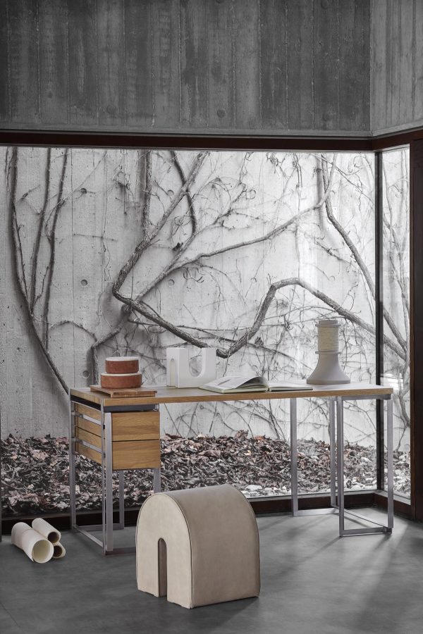 Et minimalistisk arbejdsområde med et elegant hvidt skrivebord med forskellige keramikstykker, sat op ad en stor betonvæg med et detaljeret vægmaleri af nøgne træer. En unik, buet stol fra TomorrowsDesign støder op til