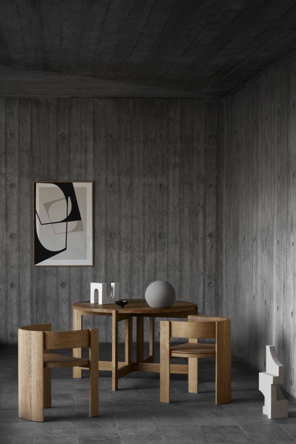 Minimalistisk spisestue med træbord og stole, sfærisk indretning og abstrakt kunst på en tekstureret betonvæg. Et lys og en lille skulptur fra TomorrowsDesign står på bordet.