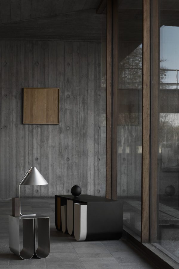 Et minimalistisk interiør med store glasvinduer, betonvægge og mørkt klinkegulv. Funktionerne omfatter en slank, metallisk skrivebordslampe, en poleret sort skrivebordsarrangør og reflekterende dekorative vaser.
