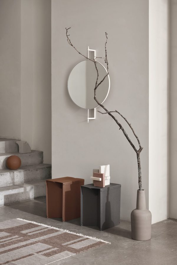 Et minimalistisk indvendigt hjørne med KRISTINA DAM - Roterende Spejl spejl på en beige væg, en stor gren i en grå vase, ved siden af hvilken der er to blokformede, kubeformede taburetter og et mønstret tæppe på gulvet.